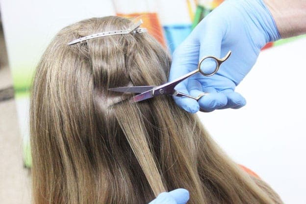 Gloved hand using silver scissors to cut a strand of long light brown hair.